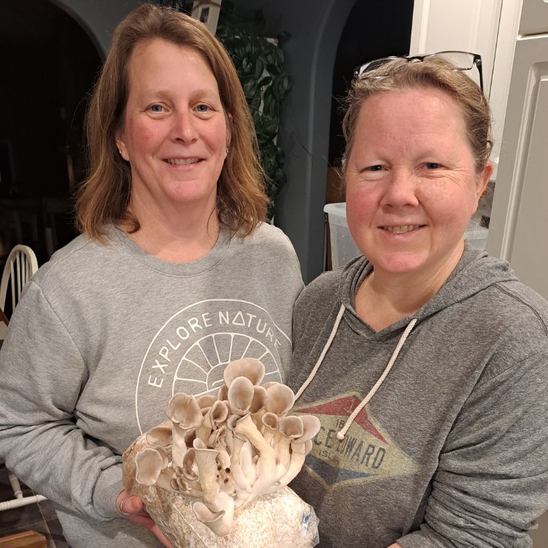 Two women hold a fruiting block of oyster mushrooms.