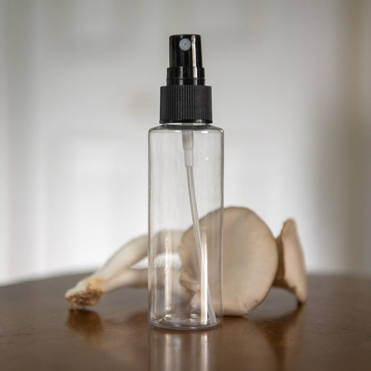 A small water mister with a black cap sits in front of two oyster mushrooms on a wooden table.